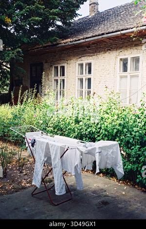 Blanchisserie blanche séchant sur une corde à linge dans un jardin, avec une vieille maison en briques et des fenêtres vintage en arrière-plan. Banque D'Images