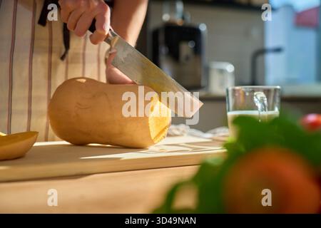 Personne dans le tablier coupant la courge butternut sur la planche de bois dans la cuisine. Gros plan de femme tranchant de la citrouille fraîche avec des légumes frais et des herbes sur la table. Banque D'Images