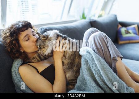 Jeune femme aux cheveux rouges embrassant un chat moelleux gris, assise sur un canapé moelleux, recouvert d'une couverture Banque D'Images