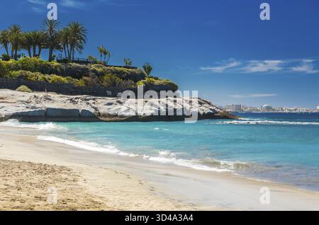 La beauté tranquille de Playa Del Duque, Tenerife, avec un rivage de sable avec des vagues douces, des formations rocheuses avec des palmiers luxuriants, et un SK bleu clair Banque D'Images