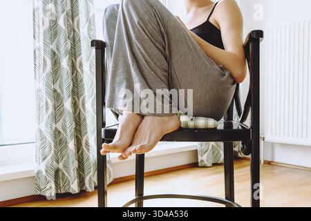 Jeune femme en pyjama, débardeur, pieds nus, assise sur une chaise en bois, jambes cachées sous elle, près d'une grande fenêtre lumineuse. concept matinal insouciant, profitez du lon Banque D'Images