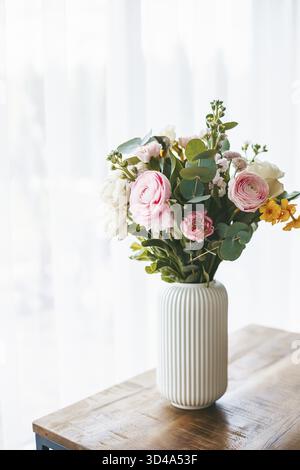 Un magnifique bouquet de fleurs de différentes teintes et types remplit un vase blanc côtelé, debout sur une table en bois Banque D'Images