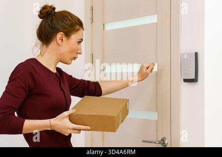 Femme tenant le paquet atteint pour le clavier sur la porte. Vue latérale d'une cliente avec une boîte en carton approchant du verrou électronique au point de ramassage Banque D'Images
