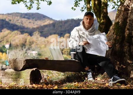 Homme présentant une feuille vide tout en travaillant sur un ordinateur portable dans une forêt d'automne pittoresque avec accompagnement de café. Présentant une feuille de papier vide, un homme travaille Banque D'Images