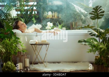 Une femme souriante aux cheveux foncés s'incline dans une baignoire blanche à bulles, les yeux fermés dans la détente. Entouré de plantes vertes luxuriantes, vapeur et produits de bain, Banque D'Images