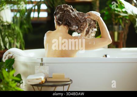 Vue de côté et de dos, une femme lave ses cheveux foncés et ondulés, recouverts de mousse blanche, dans une baignoire blanche. Ce moment privé de soins personnels et de relaxation Banque D'Images