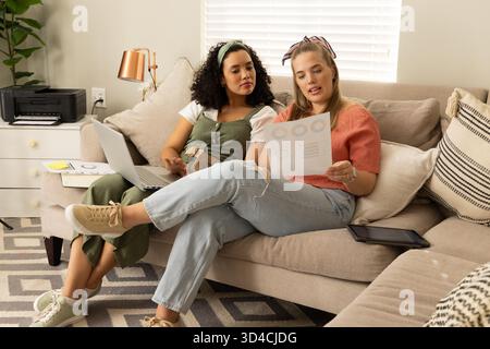 Diverses amies féminines examinant les cartes imprimées à l'aide d'un ordinateur portable sur le canapé au bureau à domicile, avec imprimante Banque D'Images