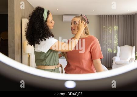 Maquilleuse féminine diversifiée et cliente appliquant le maquillage des sourcils dans la chambre à coucher, avec lumière annulaire Banque D'Images