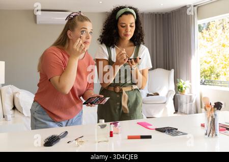 Application de maquillage des yeux diverses amies féminines debout à la vanité dans la chambre, avec des pinceaux et des palettes Banque D'Images