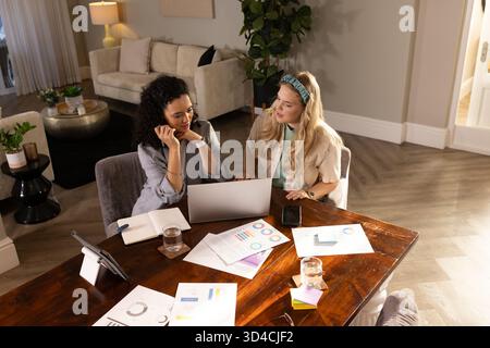 Collaborer diverses amies féminines analysant des graphiques imprimés dans un salon moderne, avec ordinateur portable Banque D'Images