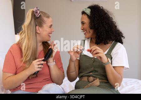 Rire diverses amies féminines se brossant les cheveux et appliquant du rouge à lèvres sur le lit blanc, avec une brosse à cheveux Banque D'Images