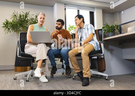 Collaboration de divers collègues assis autour d'un ordinateur portable dans un espace de bureau moderne, avec des plantes en pot Banque D'Images
