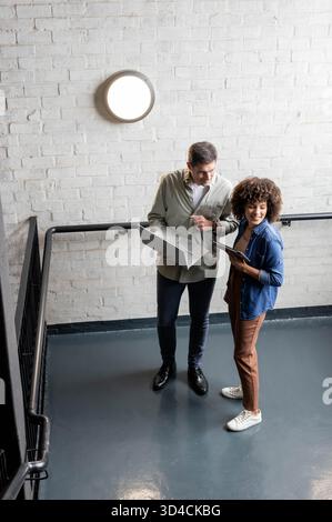 Debout divers collègues examinant la tablette et l'ordinateur portable sur la cage d'escalier du bureau, avec balustrade et lumière Banque D'Images