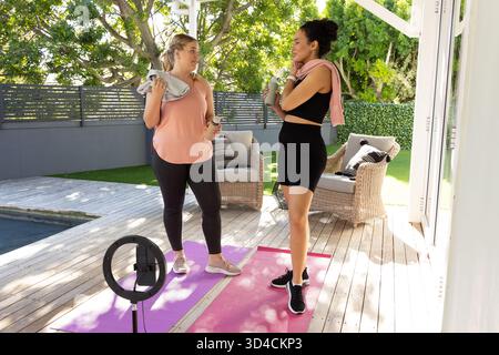 Bavardez diverses amies féminines sur le patio en bois à côté de la piscine, avec des tapis de yoga et un anneau lumineux Banque D'Images