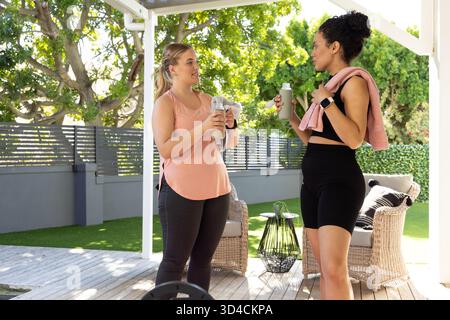 Bavarder diverses amies féminines s'hydratant sur la terrasse en bois sous la pergola, avec des bouteilles d'eau et des serviettes Banque D'Images
