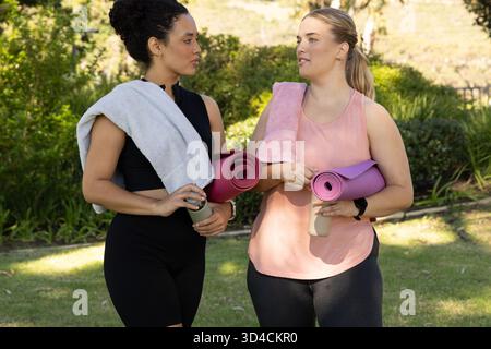 Diverses amies féminines discutant dans le parc ensoleillé, avec des tapis de yoga serviettes bouteilles d'eau Banque D'Images