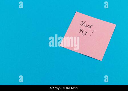 Une note autocollante rose avec un texte manuscrit disant merci est placée sur un fond bleu vif, créant un message joyeux et positif. Banque D'Images
