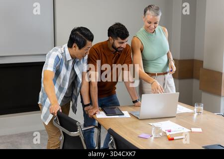 Examen de divers collègues penchés au-dessus de la table dans la salle de conférence, avec ordinateur portable et graphiques Banque D'Images