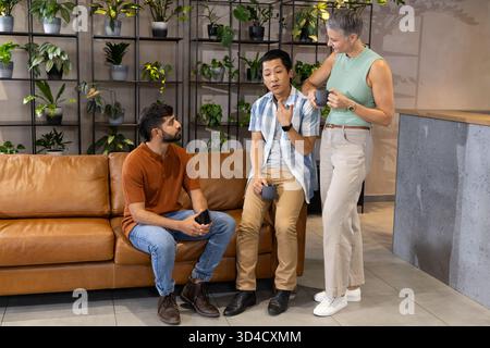 Discuter avec divers collègues assis sur le canapé dans le salon, avec mur végétal, smartphone et tasses à café Banque D'Images