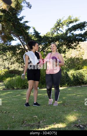 Diverses amies féminines discutant sur la pelouse herbeuse du parc, avec des tapis de yoga roulés et des bouteilles d'eau Banque D'Images