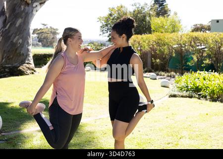 Étirer diverses amies féminines tenant les pieds derrière sur la pelouse herbeuse dans le parc, avec des lumières de jardin Banque D'Images