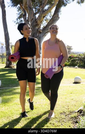 Marchant diverses amies féminines portant des tapis de yoga roses et violets dans le parc ensoleillé, avec des ornements Banque D'Images