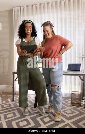 Tenant la tablette, diverses collègues femmes debout côte à côte dans le bureau à domicile, examinant le contenu Banque D'Images
