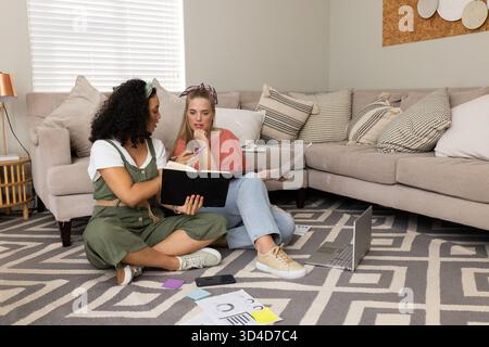 Étudiantes diverses assis les jambes croisées sur le tapis du salon, étudiant avec un ordinateur portable et des cahiers Banque D'Images