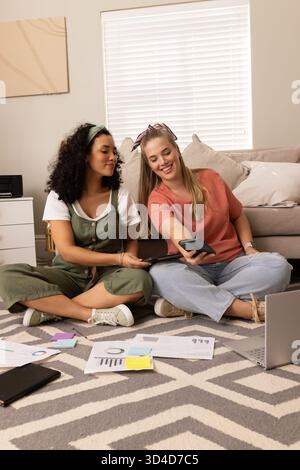 Diverses amies féminines collaborant sur un tapis chevron dans le salon, avec des cartes imprimées et un ordinateur portable Banque D'Images