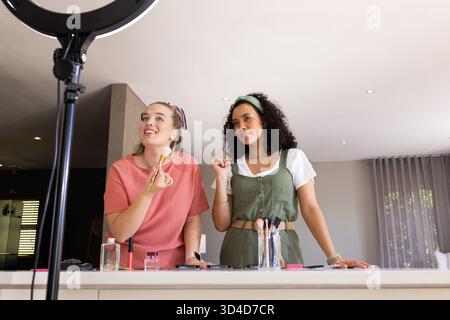 Souriant diverses amies féminines préparant le maquillage sur l'îlot de cuisine, avec lumière annulaire et brosses Banque D'Images