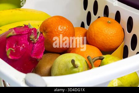 Gros plan d'un mélange vibrant de fruits frais dans un panier en plastique blanc : oranges/mandarines, bananes, fruits du dragon, kiwi et poires. Manger sainement, groce Banque D'Images