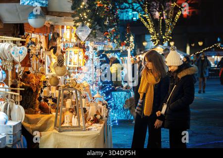Les visiteurs parcourent un étal de marché rempli d'artisanat et de décorations de vacances au marché de Noël annuel dans la soirée, à Asti, en Italie. Banque D'Images
