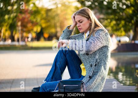 Portrait de femme d'affaires stressée en manteau vérifiant l'heure sur sa montre-bracelet tout en étant assise sur le banc du parc dans la ville. Banque D'Images