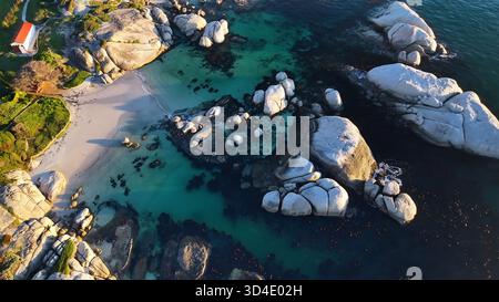 Vue aérienne de l'océan turquoise embrassant doucement les énormes rochers ensoleillés et la plage de sable près d'un bâtiment blanc solitaire, Cape Town, Wester Banque D'Images