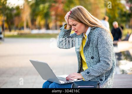 : Portrait d'une femme d'affaires surchargée et stressée avec mal de tête en utilisant un ordinateur portable assis sur un banc de parc dans la ville. Banque D'Images