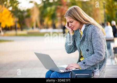 Portrait de femme d'affaires surchargée et stressée avec mal de tête en utilisant un ordinateur portable tout en étant assis sur un banc de parc dans la ville. Banque D'Images