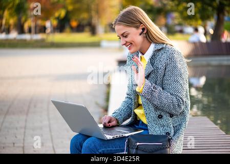 Portrait de femme d'affaires en manteau portant des écouteurs et agitant pendant un appel vidéo sur ordinateur portable tout en étant assis sur le banc du parc dans la ville. Banque D'Images