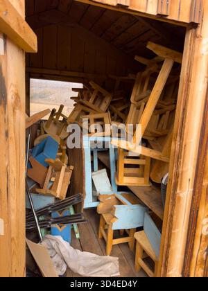 Scène chaotique à l'intérieur d'un hangar en bois emballé avec des chaises, des tables et des planches empilées de manière désordonnée. Les meubles bleus altérés contrastent les tons chauds du bois créant une ambiance de rangement rustique. Banque D'Images