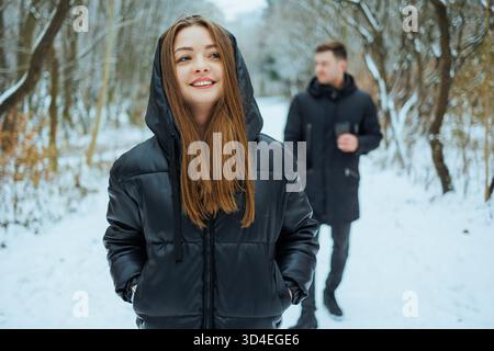 Par un joyeux jour d'hiver, une jeune femme joyeuse dans un élégant manteau noir profite de sa promenade le long d'un chemin enneigé, avec son compagnon se promenant derrière elle Banque D'Images