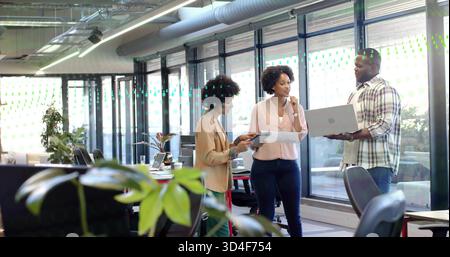 Discuter avec des collègues en tenue professionnelle partageant ordinateur portable, tablette et documents dans un bureau moderne Banque D'Images