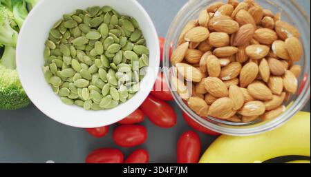 Affichage de deux bols de pepitas et d'amandes sur le comptoir gris foncé avec tomates, banane, brocoli Banque D'Images