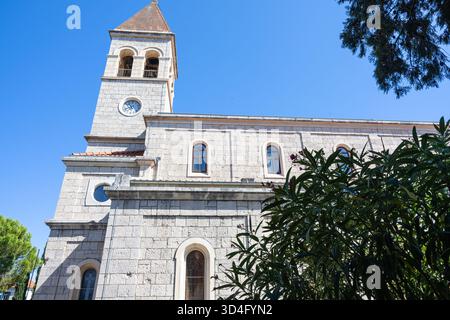 Une église historique en pierre dispose d'une grande tour de l'horloge, de fenêtres cintrées et d'un toit en tuiles. Ensemble contre un ciel bleu vif avec un arbre luxuriant, mettant en évidence Ar Banque D'Images