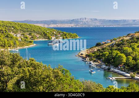 Une crique méditerranéenne tranquille avec de l'eau turquoise, des bateaux amarrés et une rive bordée d'arbres. Collines verdoyantes et petites maisons le long de la côte créent un Banque D'Images