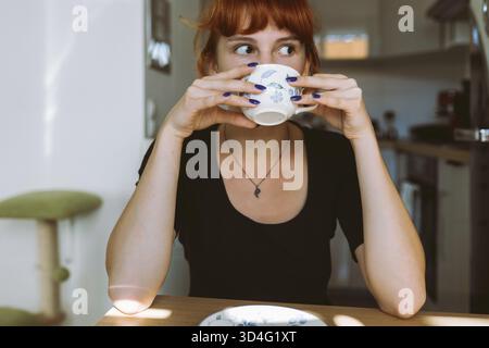 Portrait jeune femme aux cheveux roux attachés dans un pain assis à table, tenant tasse et soucoupe dans les mains, buvant du thé vert, infusion de plantes, café, matin Banque D'Images