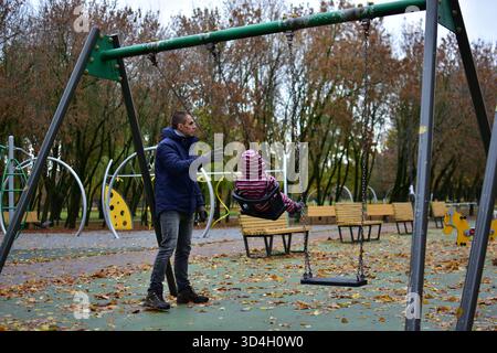 Adulte poussant le tout-petit sur la balançoire dans le terrain de jeu pendant la journée froide d'automne, entouré d'arbres et de feuilles colorées. Doux lien familial et activ en plein air Banque D'Images
