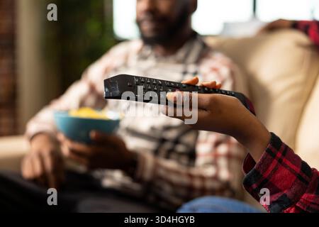 Gros plan sur la main de la femme noire tenant la télécommande, visant à la télévision comme fond montre l'homme tenant le bol de collations. Afro-américain utilisant un appareil pour retourner à travers les chaînes de télévision. Banque D'Images