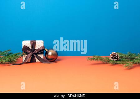 Boîte cadeau avec ruban marron, branches de sapin, pomme de pin et boule sur fond bleu et orange. Carte de voeux joyeux Noël, espace de copie Banque D'Images
