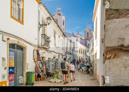 Lagos, Portugal, le 8 novembre 2025, les visiteurs se promènent dans une rue étroite animée remplie de cafés, de stands de marché et de vues sur une église historique. Banque D'Images