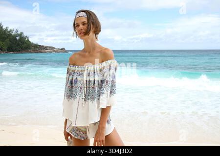Belle jeune femme en tenue d'été debout sur la plage, derrière l'océan Indien bleu turquoise, tout en tenant ses chaussures et en regardant loin. Banque D'Images