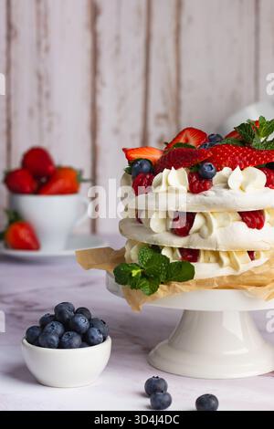 Un pavlova coloré est assis sur un stand de gâteau blanc, orné de fraises sucrées, framboises et bleuets. En arrière-plan, bols de fruits frais Banque D'Images